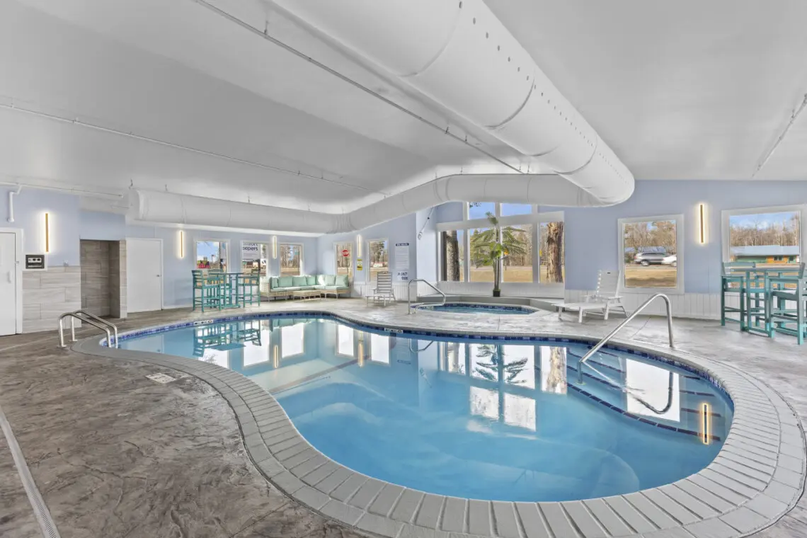 Indoor swimming pool at Nisswa Hotel & Suites in Nisswa, Minnesota. The bright, modern pool area features a large rectangular pool with clear blue water, surrounded by white lounge chairs, large windows, and modern lighting.