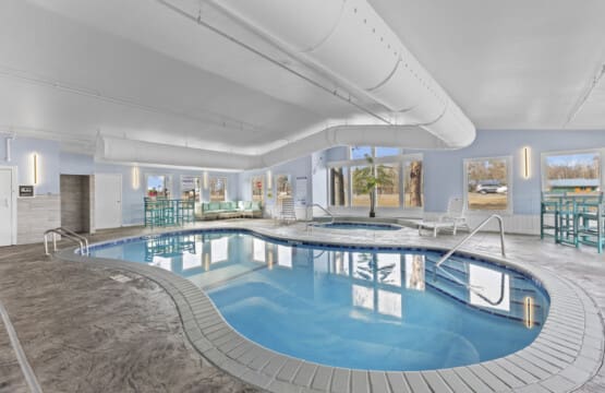 Indoor swimming pool at Nisswa Hotel & Suites in Nisswa, Minnesota. The bright, modern pool area features a large rectangular pool with clear blue water, surrounded by white lounge chairs, large windows, and modern lighting.